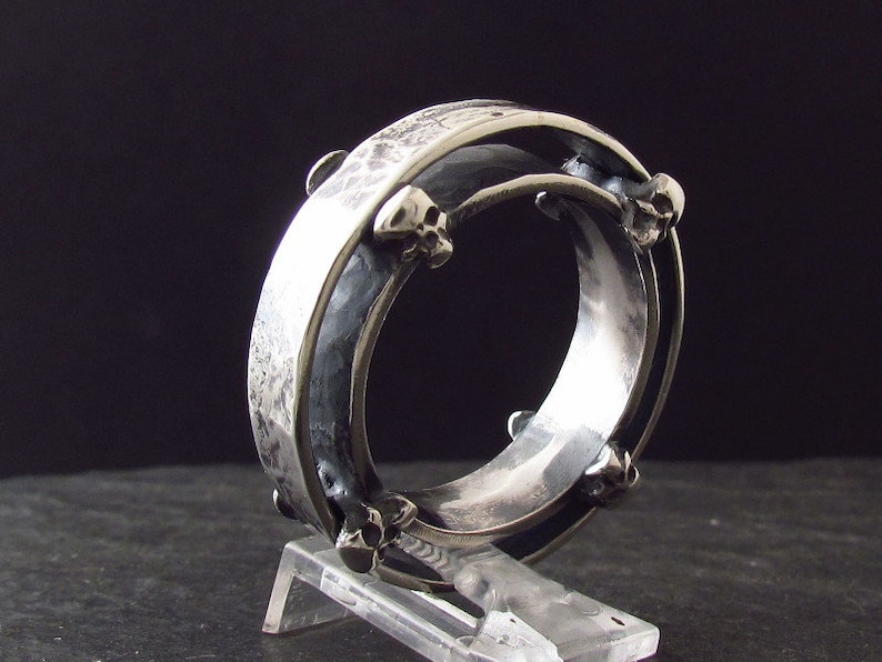 May include: A silver ring with a textured band and skull-shaped accents. The ring is made of two bands that are joined together.