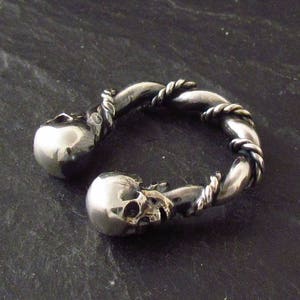 May include: A silver ring with two skull designs. The ring is twisted and has a rope-like design.