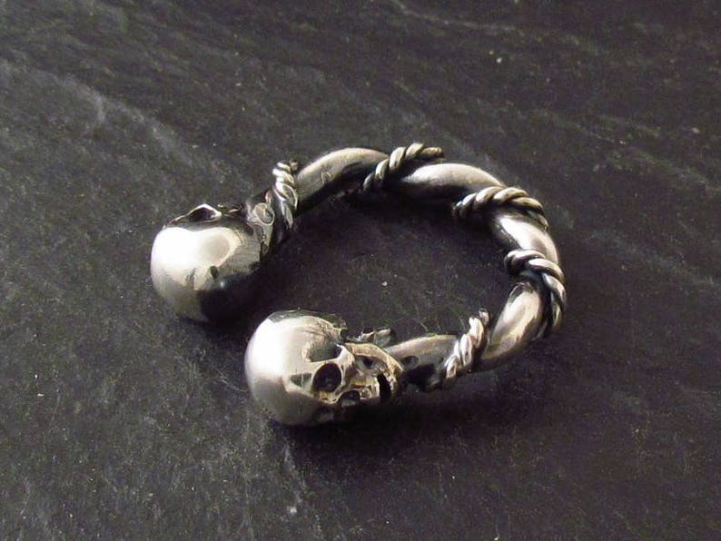 May include: A silver ring with two skull designs. The ring is twisted and has a rope-like design.