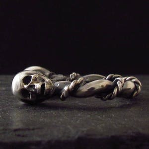 May include: A silver ring with two intertwined skull designs. The ring is made of twisted metal and has a detailed skull design.