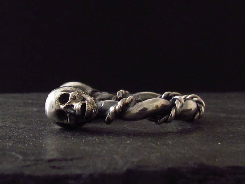 May include: A silver ring with two intertwined skull designs. The ring is made of twisted metal and has a detailed skull design.