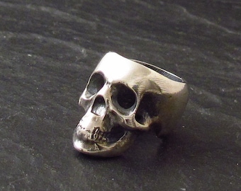 May include: A silver skull ring with a detailed design. The ring is made of metal and has a smooth finish.