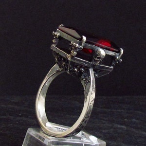 May include: A silver ring featuring a large, dark red gemstone set within an intricate, skeletal design.