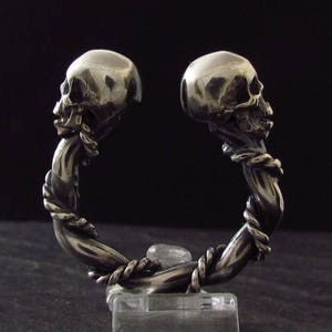 May include: A silver bracelet with two skull heads facing each other. The bracelet is made of a twisted metal design.