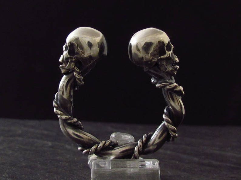 May include: A silver bracelet with two skull heads facing each other. The bracelet is made of a twisted metal design.