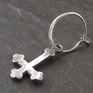 May include: A silver hoop earring with a silver cross charm. The cross has a textured surface and is in the shape of a traditional cross.