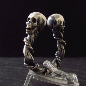 May include: A silver skull ring with two skulls intertwined, one facing forward and the other facing backward. The ring is made of twisted metal and has a detailed design.