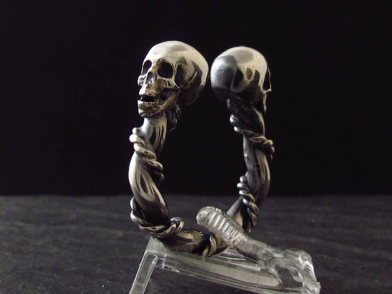 May include: A silver skull ring with two skulls intertwined, one facing forward and the other facing backward. The ring is made of twisted metal and has a detailed design.