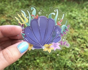 Puede incluir: Una pegatina transparente con una concha de mar púrpura, flores amarillas y rosas y un tridente. La pegatina está sobre un fondo verde.