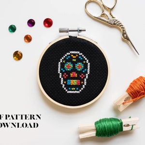 May include: A colourful sugar skull cross stitch pattern in a black embroidery hoop. The hoop is surrounded by colourful rhinestones, embroidery floss, and embroidery scissors. The text "PDF PATTERN DOWNLOAD" is at the bottom of the image.