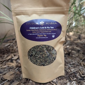 May include: A brown paper bag with a clear window showing a blend of dried herbs. The bag has a label that reads "DIVINA ANIMA Children's Cold & Flu Tea" and lists the ingredients: Catnip, Elderflower, Elderberry, Echinacea Root, Anise Seed, Peppermint Leaf. The label also states "Organic 70g" and provides a website address: www.divinaanima.com.