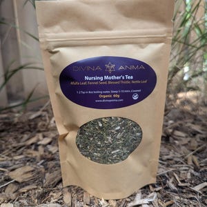 Op de afbeelding: Een bruine papieren zak met een paars en wit etiket waarop staat "DIVINA ANIMA Nursing Mother's Tea Alfalfa Leaf, Fennel Seed, Blessed Thistle, Nettle Leaf 1-2 Tsp in 8oz boiling water, Steep 5-10 mins Covered Organic 60g www.divinaanima.com". De zak bevat losse groene theebladeren.
