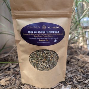 Puede incluir: Una bolsa de papel marrón con una ventana transparente que muestra una mezcla de hierbas secas. La bolsa está etiquetada como "Third Eye Chakra Herbal Blend" con una lista de ingredientes que incluye manzanilla, hoja de arándano, skullcap, raíz de valeriana, hoja de ortiga, flor de saúco y flor de la pasión. La bolsa también indica "Organic 50g" e incluye una dirección de sitio web.