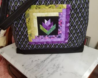 May include: A handmade tote bag with a black base and straps. The bag features a patchwork design with a central tulip motif in purple, green, and pink. The main fabric has a black background with a repeating pattern of green leaves.