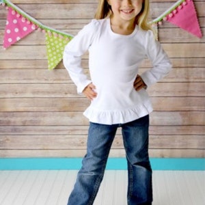 Puede incluir: Una ni&ntilde;a peque&ntilde;a con una camisa blanca de manga larga con dobladillo con volantes y vaqueros azules. Tiene un gran lazo blanco en el pelo y est&aacute; de pie frente a un fondo de madera con banderines de colores.