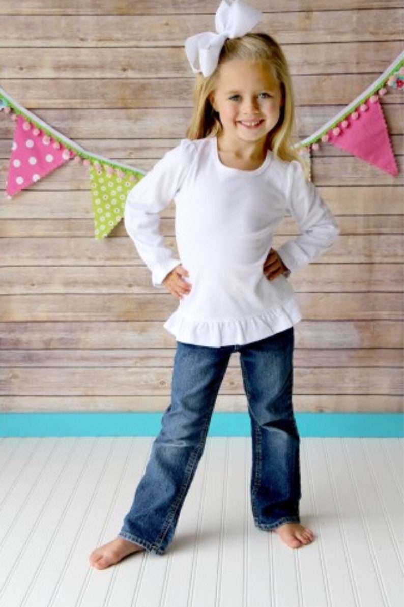 Puede incluir: Una ni&ntilde;a peque&ntilde;a con una camisa blanca de manga larga con dobladillo con volantes y vaqueros azules. Tiene un gran lazo blanco en el pelo y est&aacute; de pie frente a un fondo de madera con banderines de colores.