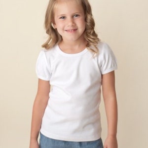 Puede incluir: Una ni&ntilde;a peque&ntilde;a con una camiseta blanca de manga corta abullonada y pantalones cortos vaqueros. La camiseta tiene cuello redondo. Los pantalones cortos son de mezclilla azul claro. La ni&ntilde;a sonr&iacute;e.