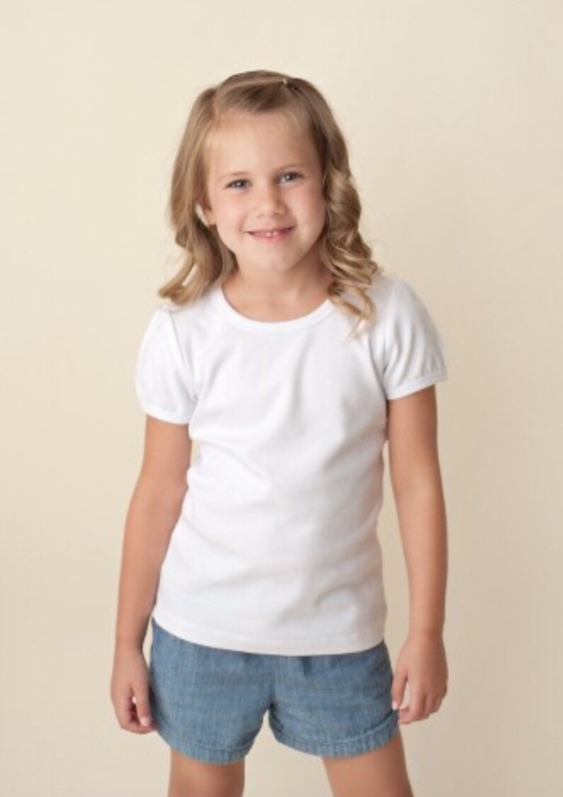 Puede incluir: Una ni&ntilde;a peque&ntilde;a con una camiseta blanca de manga corta abullonada y pantalones cortos vaqueros. La camiseta tiene cuello redondo. Los pantalones cortos son de mezclilla azul claro. La ni&ntilde;a sonr&iacute;e.