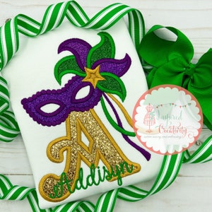 Puede incluir: Camiseta blanca con un dise&ntilde;o de m&aacute;scara de Mardi Gras. La m&aacute;scara es morada con detalles verdes y dorados, y una letra "A" dorada con purpurina con el nombre "Addisyn" en escritura verde. Cinta a rayas verdes y blancas y un lazo verde en el fondo.