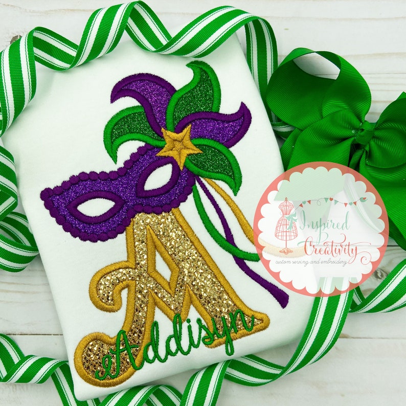 Puede incluir: Camiseta blanca con un dise&ntilde;o de m&aacute;scara de Mardi Gras. La m&aacute;scara es morada con detalles verdes y dorados, y una letra "A" dorada con purpurina con el nombre "Addisyn" en escritura verde. Cinta a rayas verdes y blancas y un lazo verde en el fondo.
