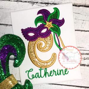 Puede incluir: Tela blanca con un dise&ntilde;o de Mardi Gras. Presenta una m&aacute;scara morada, detalles florales verdes y morados, y una letra "C" dorada con purpurina. El nombre "Catherine" est&aacute; bordado en verde.
