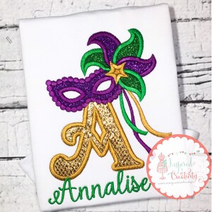 Puede incluir: Camiseta blanca con un dise&ntilde;o de Mardi Gras. El dise&ntilde;o presenta una m&aacute;scara de purpurina morada, plumas de purpurina verdes y moradas, una letra "A" dorada con purpurina, una estrella dorada y el nombre "Annalise" en escritura verde.