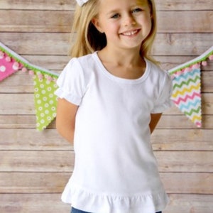 Puede incluir: Una ni&ntilde;a peque&ntilde;a con una camiseta blanca de manga corta con dobladillo con volantes y vaqueros azules. Tiene un gran lazo blanco en el pelo rubio. Una colorida pancarta con pompones cuelga en el fondo. El fondo es una pared de madera marr&oacute;n claro.