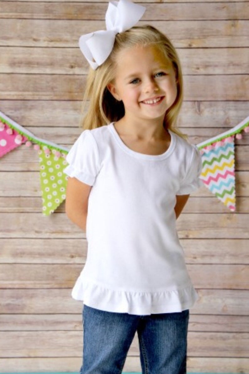 Puede incluir: Una ni&ntilde;a peque&ntilde;a con una camiseta blanca de manga corta con dobladillo con volantes y vaqueros azules. Tiene un gran lazo blanco en el pelo rubio. Una colorida pancarta con pompones cuelga en el fondo. El fondo es una pared de madera marr&oacute;n claro.