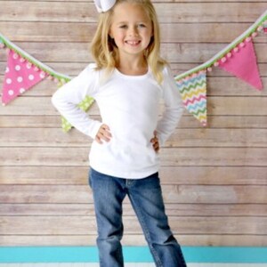 Puede incluir: Una ni&ntilde;a con una camiseta blanca de manga larga y vaqueros azules, con un gran lazo blanco en el pelo. El fondo presenta una pared de madera y banderines coloridos.