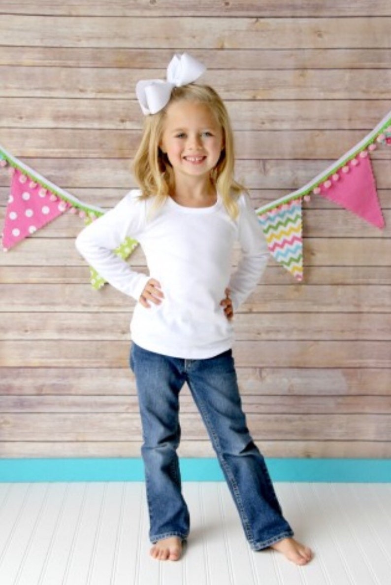 Puede incluir: Una ni&ntilde;a con una camiseta blanca de manga larga y vaqueros azules, con un gran lazo blanco en el pelo. El fondo presenta una pared de madera y banderines coloridos.