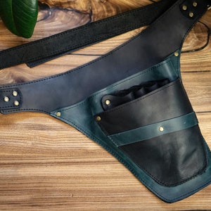 Pode incluir: Um aljava de couro em tons de preto, verde-azulado e azul marinho, com uma al&ccedil;a de ombro e uma bolsa. A aljava foi projetada para ser usada na transversal. O couro &eacute; costurado e rebitado, e o fundo &eacute; uma superf&iacute;cie de madeira.