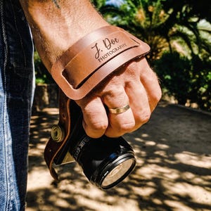 May include: A tan leather camera hand strap featuring a wide, curved wrist pad with the engraved text J. Doe PHOTOGRAPHY. The strap is attached to a black camera lens and body, designed to provide a secure grip while holding the camera device.