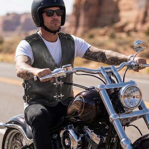 May include: A dark olive green leather vest featuring a V-neckline, silver-toned metal buckles on the sides, and decorative silver studs along the shoulders and front closure, worn over a plain white short-sleeved t-shirt.