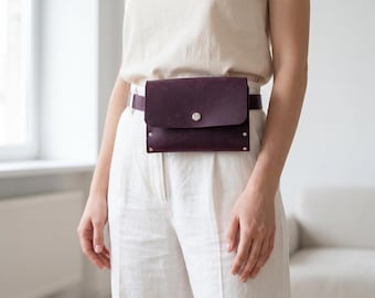 May include: A burgundy leather fanny pack with a flap closure and silver-tone hardware. The belt bag is worn around the waist over white pants and a cream-colored top. The bag has a minimalist design.