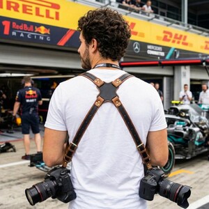 May include: A brown and black leather camera harness with two black cameras suspended from it. The harness is worn by a person in a white t-shirt. The background shows a Formula 1 racing event.