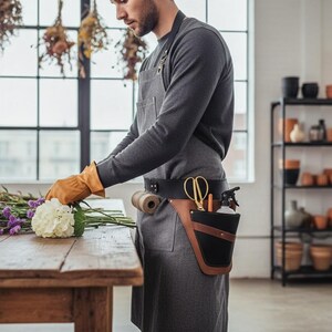 Pode incluir: Uma pessoa usando um avental cinza e luvas bege arrumando flores em uma mesa de madeira. Um cinto de ferramentas de couro preto e marrom com tesouras douradas e um borrifador &eacute; vis&iacute;vel. Flores secas penduradas no fundo.