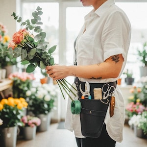 Pode incluir: Um florista segurando um buqu&ecirc; de rosas cor-de-rosa e eucalipto. O florista usa uma camisa branca e um cinto de ferramentas de couro preto com tesoura, barbante e uma faca. O fundo mostra uma floricultura.