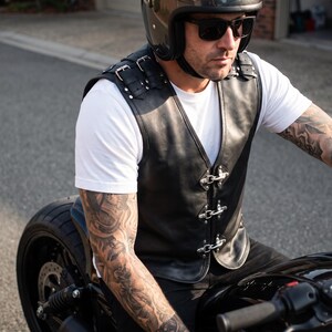 May include: A black leather vest featuring silver-toned metal buckles on the shoulders and three silver-toned metal hook-and-eye closures down the front. The vest is worn over a plain white t-shirt and includes decorative silver studs on the shoulder straps.