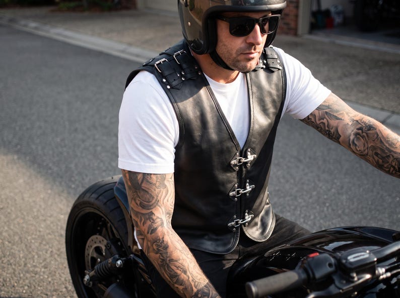 May include: A black leather vest featuring silver-toned metal buckles on the shoulders and three silver-toned metal hook-and-eye closures down the front. The vest is worn over a plain white t-shirt and includes decorative silver studs on the shoulder straps.