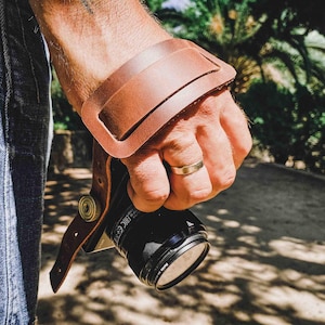 May include: A brown leather camera wrist strap with a brass buckle and a metal ring. The strap is attached to a black camera.