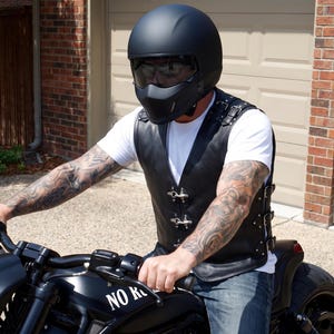 May include: A matte black motorcycle helmet with a tinted visor and a black leather vest featuring silver metal buckles, side adjustment straps, and a white t-shirt underneath. The vest is worn while operating a black motorcycle with the text NO RIDE.