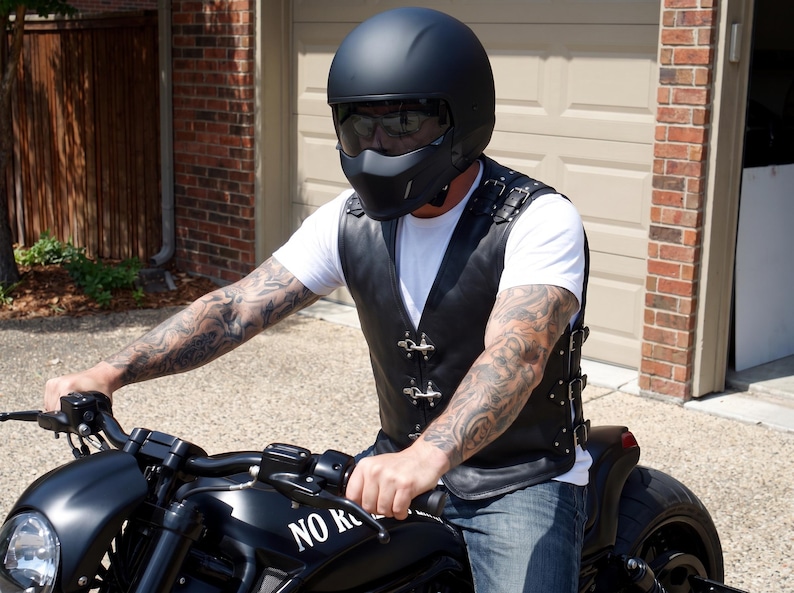 May include: A matte black motorcycle helmet with a tinted visor and a black leather vest featuring silver metal buckles, side adjustment straps, and a white t-shirt underneath. The vest is worn while operating a black motorcycle with the text NO RIDE.