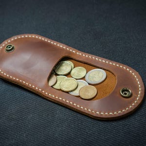 May include: A brown leather coin purse with a rectangular shape and an oval opening. The purse is filled with various coins. The edges are stitched with white thread and secured with metal rivets.