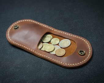 May include: A brown leather coin purse with a rectangular shape and an oval opening. The purse is filled with various coins. The edges are stitched with white thread and secured with metal rivets.
