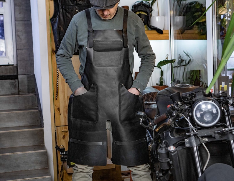 Puede incluir: Un delantal de cuero negro con m&uacute;ltiples bolsillos y correas. El delantal lo lleva puesto una persona de pie delante de una motocicleta.