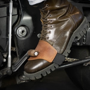 Può includere: Un proteggi-scarpa per cambio moto in pelle marrone con un inserto in pelle color cuoio liscio sulla parte superiore, fissato con un cinturino elastico nero, indossato sopra uno stivale stringato in pelle marrone scuro sul pedale del cambio.