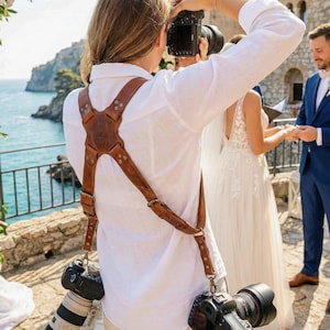 May include: A brown leather dual camera harness worn over a white long-sleeved shirt. The harness features a cross-back design with metal rivets and adjustable buckles, supporting two professional DSLR cameras with attached lenses at the hips.
