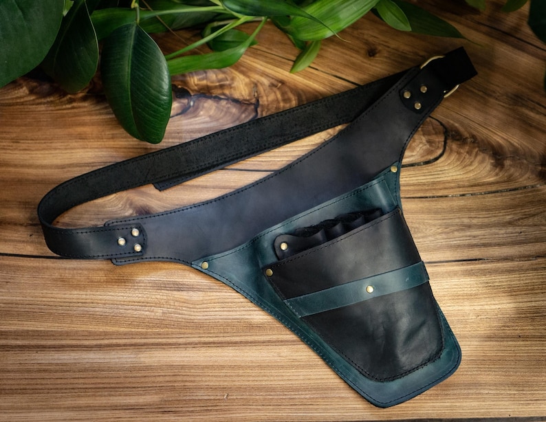Pode incluir: Um aljava de couro em tons de preto, verde-azulado e azul marinho, com uma al&ccedil;a de ombro e uma bolsa. A aljava foi projetada para ser usada na transversal. O couro &eacute; costurado e rebitado, e o fundo &eacute; uma superf&iacute;cie de madeira.