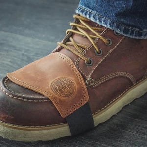 May include: A brown leather motorcycle gear shifter guard worn over a brown leather boot with tan laces and a white sole. The guard features a circular embossed logo with the text Fashion Racing and is secured with a black elastic strap.
