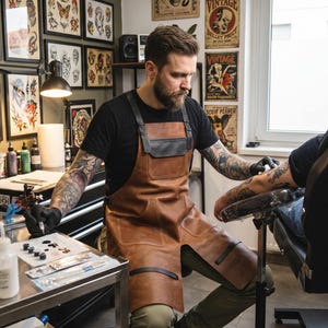 Könnte beinhalten: Eine braune Lederschürze mit schwarzen Riemen, einer Brusttasche und zwei unteren Taschen mit weißen Nähten. Die Schürze wird über einem schwarzen T-Shirt und einer olivgrünen Hose getragen und hat schwarze Akzente im unteren Bereich sowie ein schwarzes Brustteil.